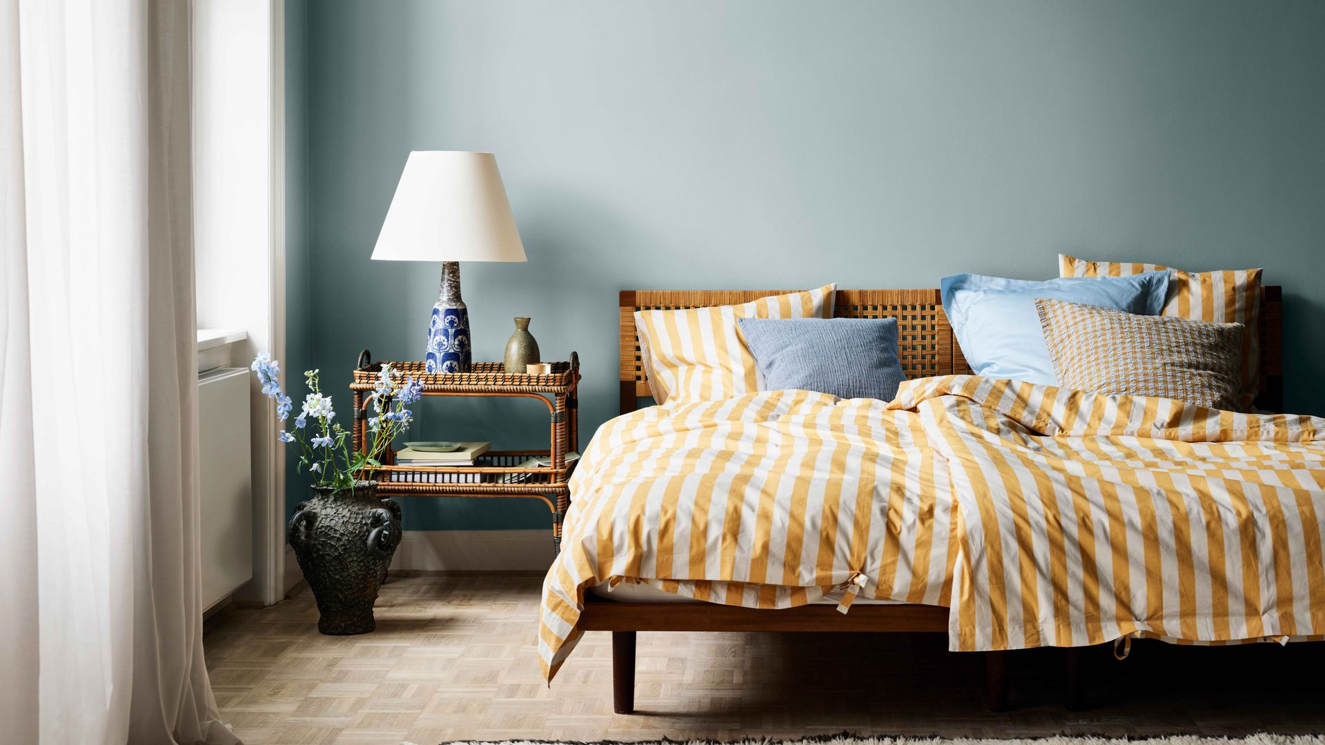 Dormitorio con paredes en azul claro, ropa de cama de rayas amarillas y blancas, cabecero de rejilla, mesita de noche de fibras vegetales, jarrón en el suelo con flores, lámpara de sobremesa con pantalla blanca y pie azul