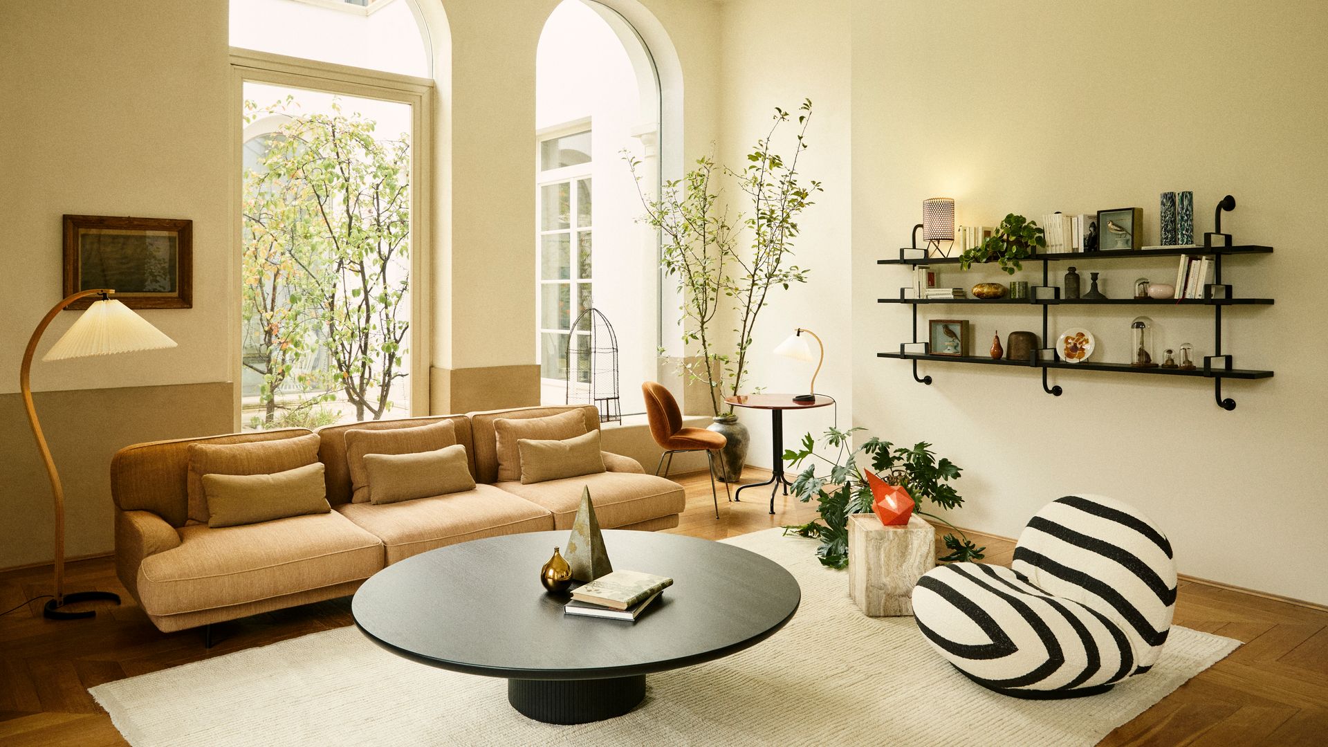 Salón con sofá marrón, mesa de centro redonda de color negro, cojines a juego, alfombra, butaca de rayas blancas y negras, estantería de pared negra y lámpara de pie