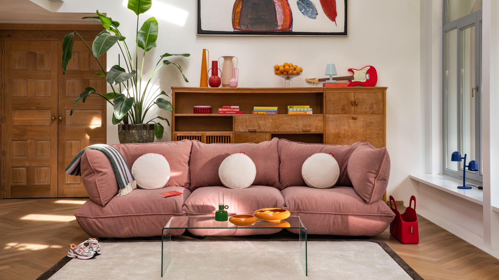 Salón con sofá rosa , planta XL, mueble de madera con baldas y cajones, mesa de plástico transparente, alfombra, cuadro en la pared 