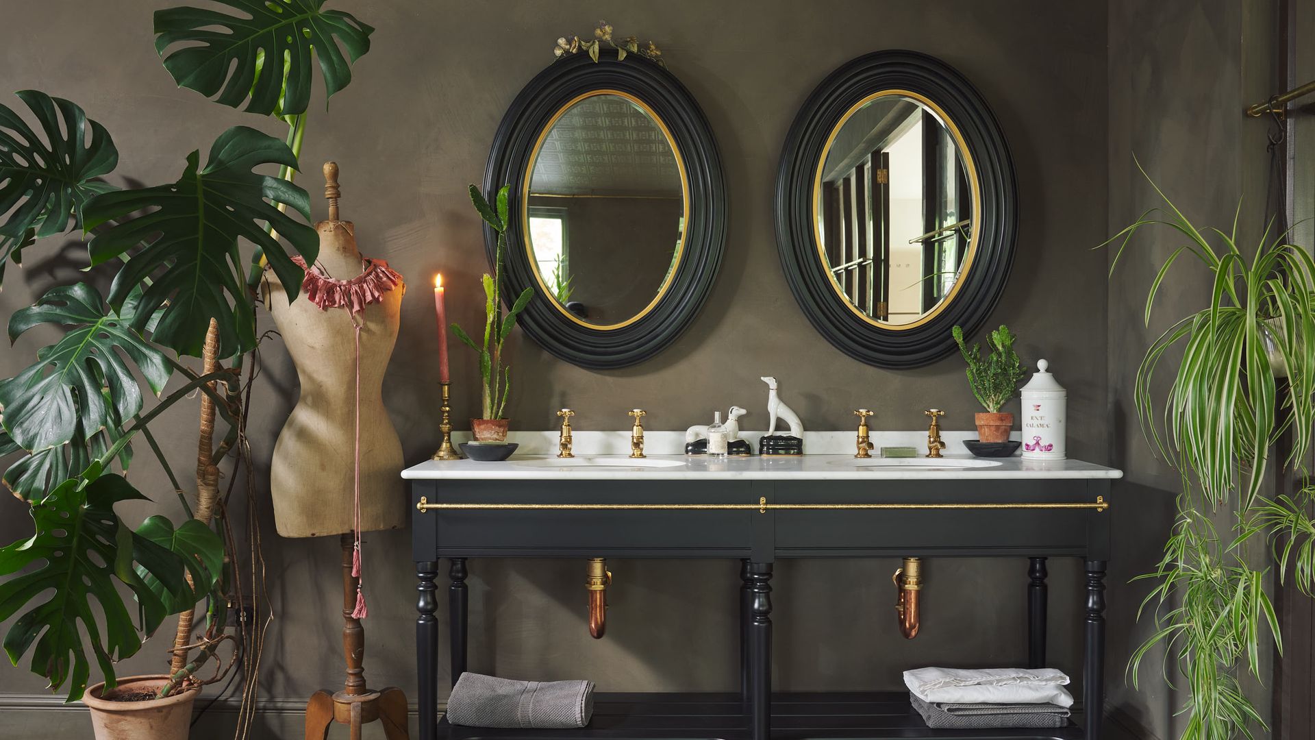 Baño con espejos ovalados, paredes en verde oscuro, espejo de baño negro con encimera blanca, plantas en el suelo, grifos dorados, adornos 