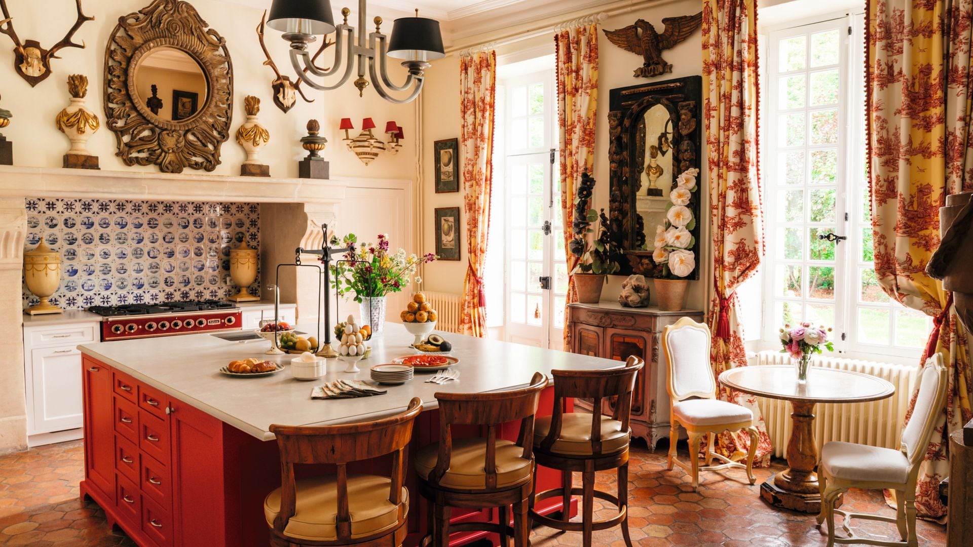 Cocina con isla roja, sillas de madera, y ventanas con cortinas estampadas.