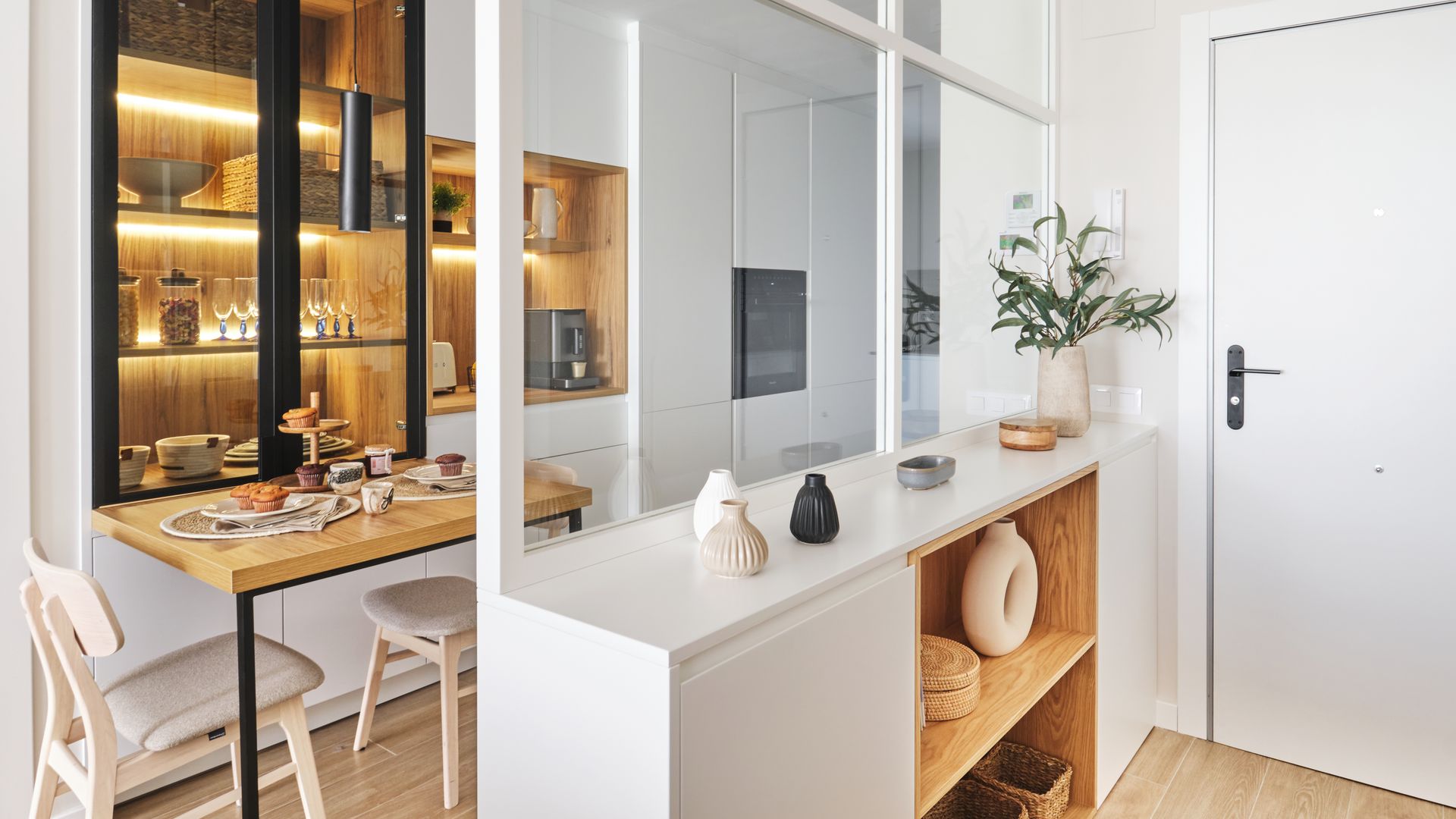 Casa pequeña con recibidor abierto con mueble, tabique de cristal separando la cocina, mesa con dos sillas, vitrina con luz 
