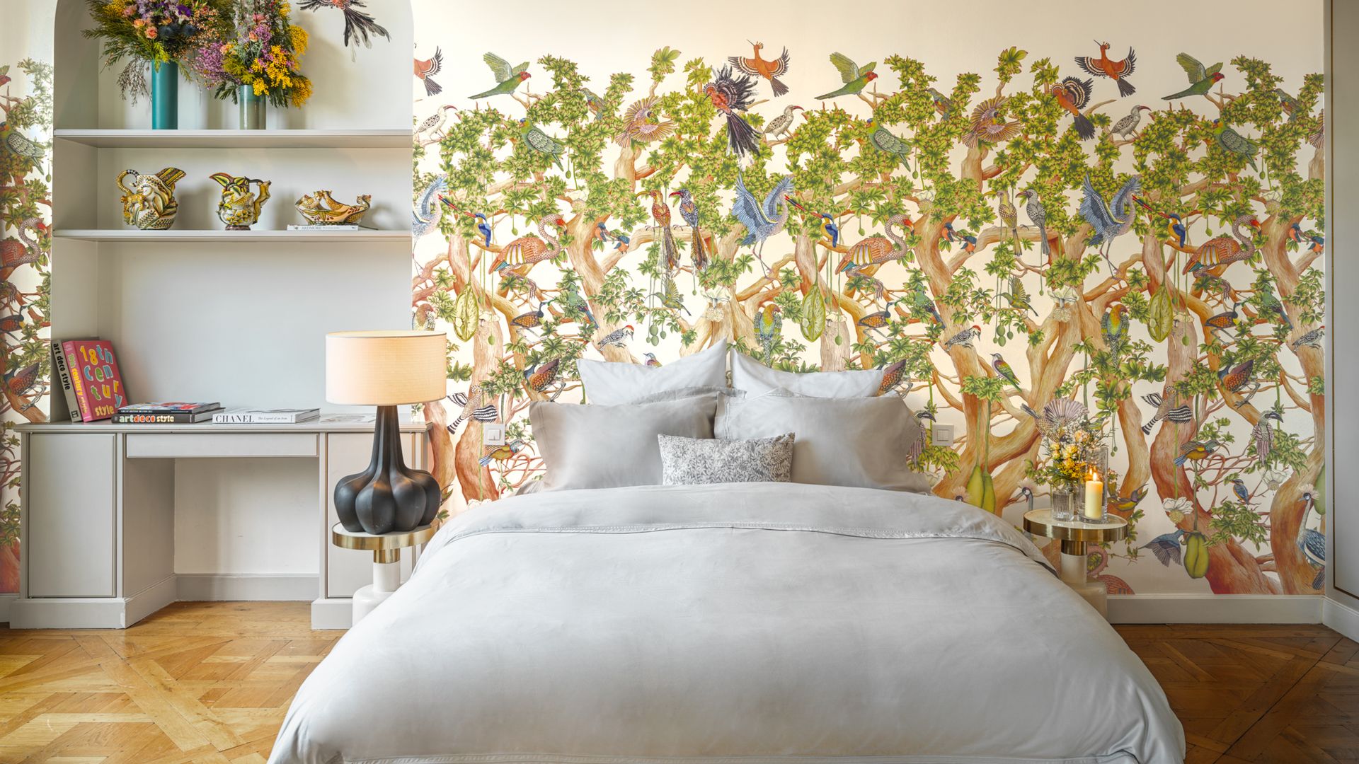 Dormitorio con papel pintado con estampado selva con pájaros y árboles, ropa de cama blanca y lámpara de sobremesa negra 