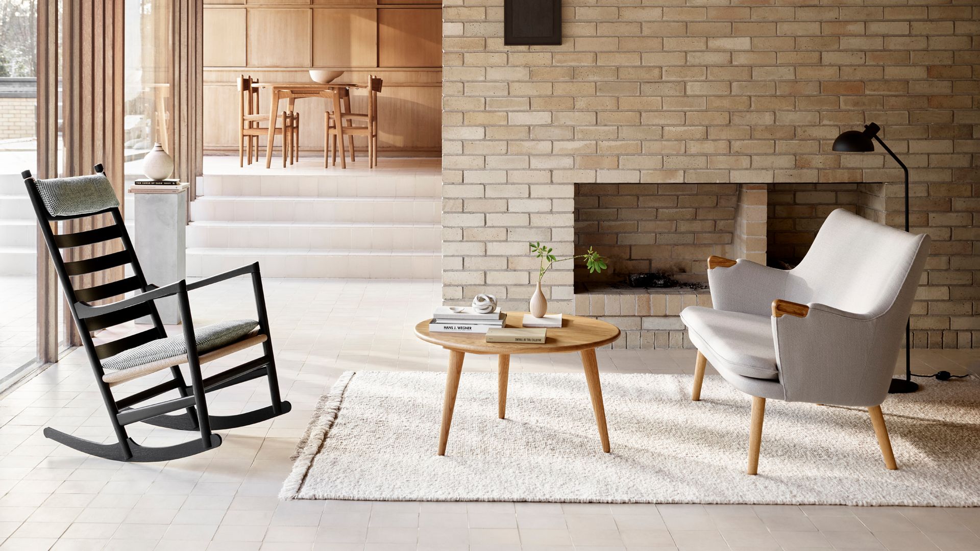 Salón con chimenea, pared de ladrillos, vigas en el techo, techo de madera, alfombra blanca, mecedora negra, mesa de centro de madera, butaca tapizada blanca