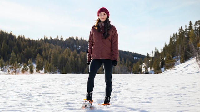 Mujer en la montaña nevada con leggins negros, cazadora acolchada y gorro granate