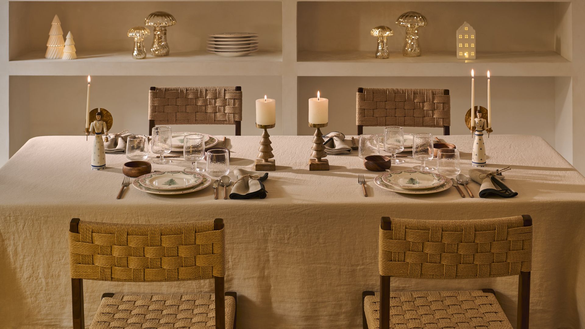 Mesa de Navidad con mantel de lino beige, velas y candelabros, sillas de comedor de fibras vegetales, estantería de obra con adornos, vajilla y corona de Navidad