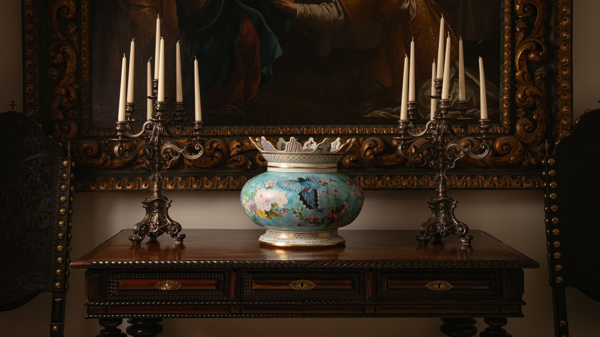 Consola antigua de madera oscura, candelabros de plata envejecida, cuadro en la pared con marco dorado y pieza de porcelana en azul 