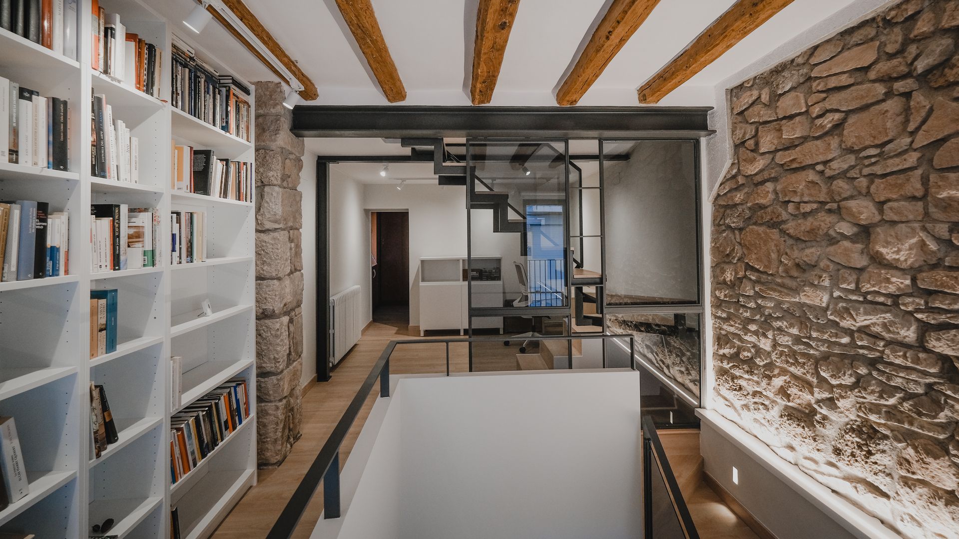 Pasillo con paredes de piedra y barandilla, vigas de madera, estantería de suelo a techo blanca con libros