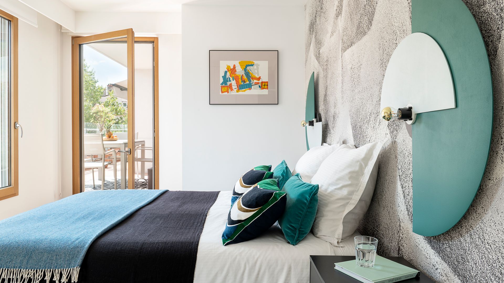 Dormitorio principal con pared del cabecero con paneles, salida a la terraza, ventana de madera, ropa de cama azul, cojines azules sobre la cama, cuadro en la pared