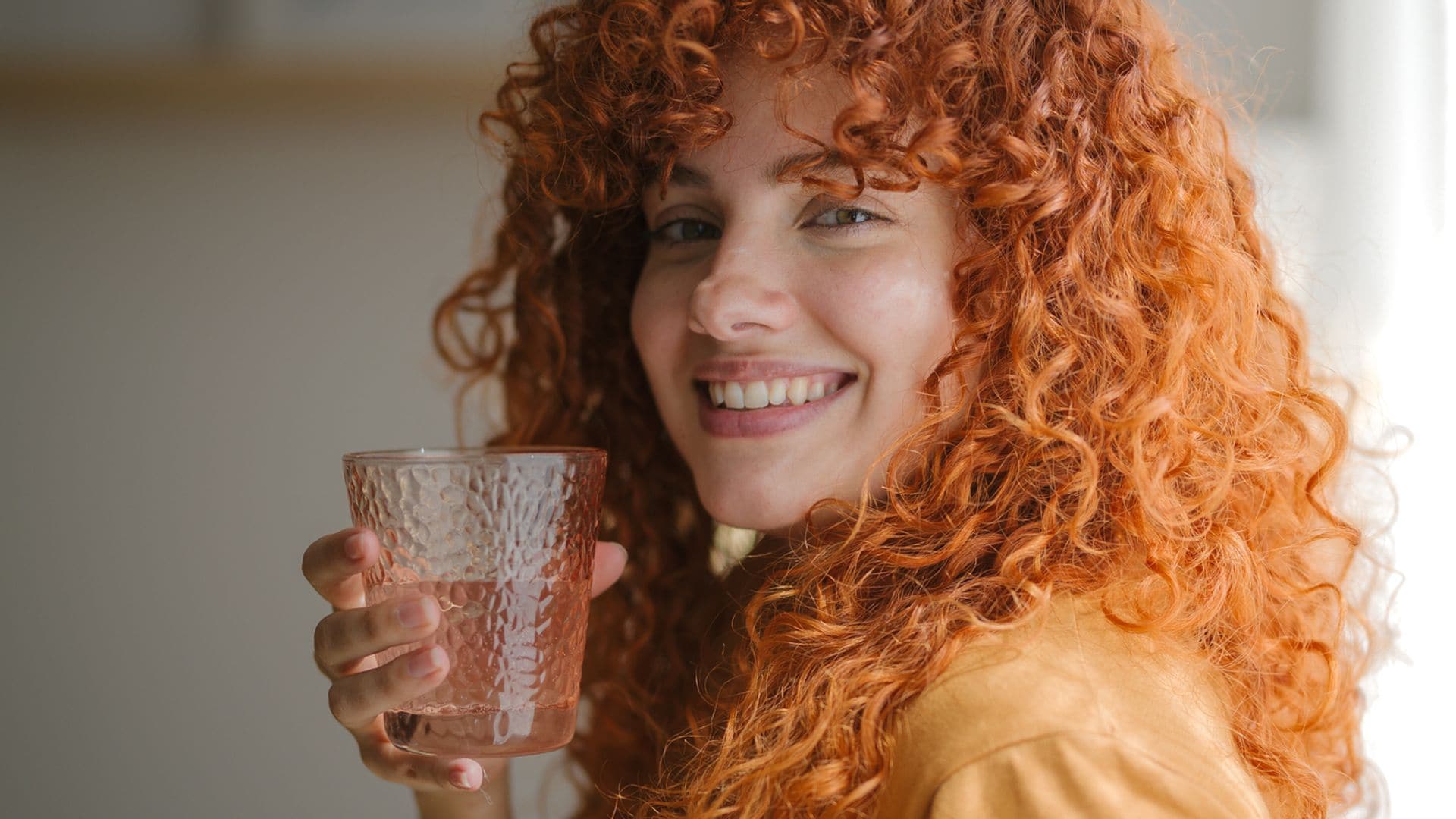 Chica joven con pelo rizado pelirrojo, sonriendo a la cámara en primer plano, con un vaso de agua en la mano y una camiseta naranja. Está apoyada sobre una mesa en casa.