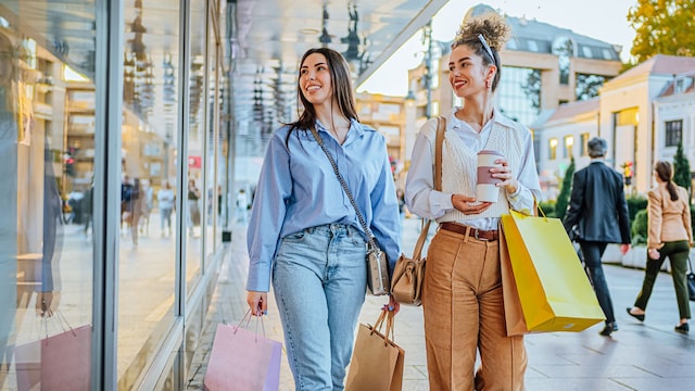 Dos chicas jóvenes sonrientes de compras con bolsas mirando escaparates desde la calle