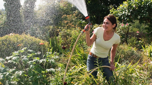 Mujer en medio de su jardín regando con una manguera hacia arriba mientras sonríe