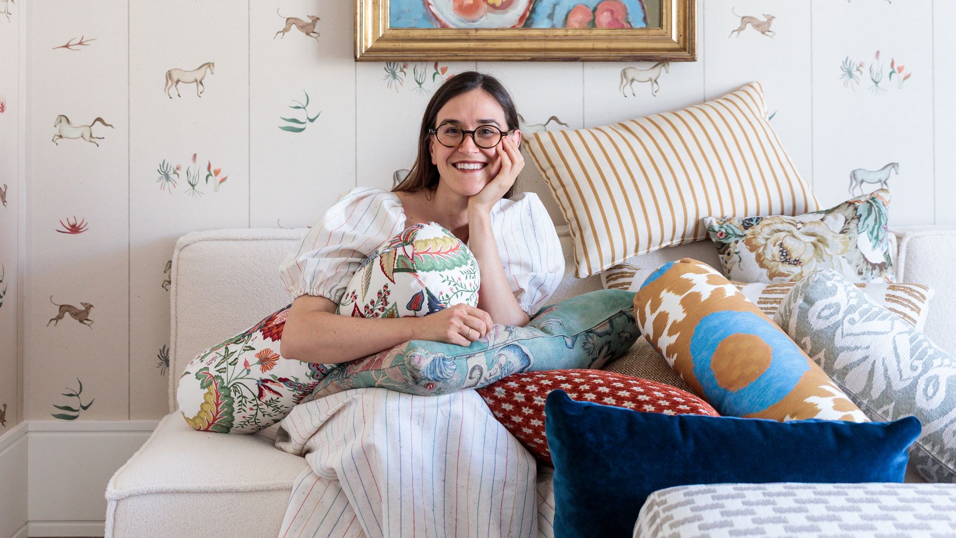 La arquitecta Pilar García-Ferrer sentada en sofá blanco, rodeada de cojines, con pared con papel pintado, cuadro con marco dorado