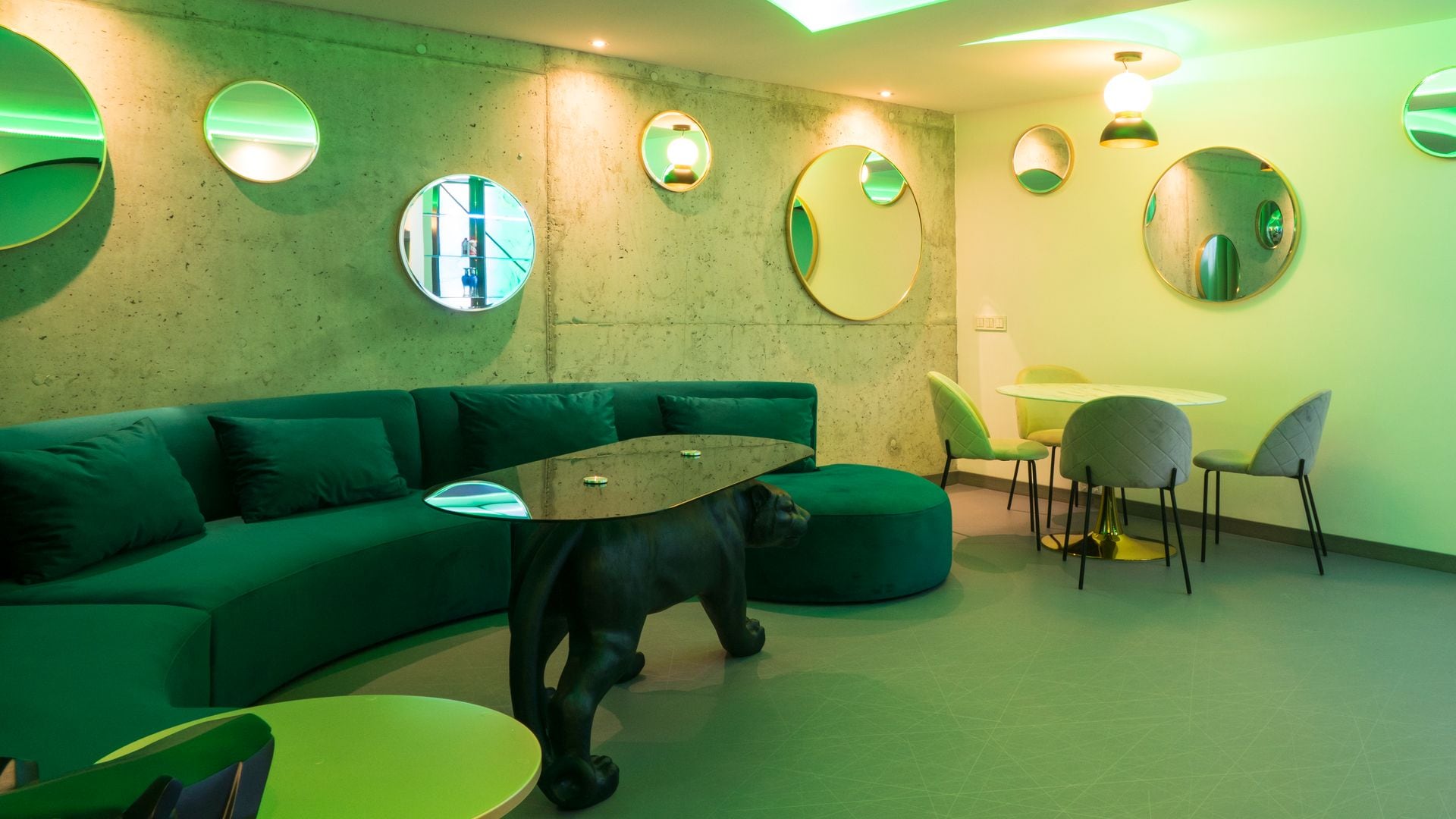 Sótano con luz de color verde, espejos, sofá curvo en verde oscuro, mesa de comedor redonda y sillas