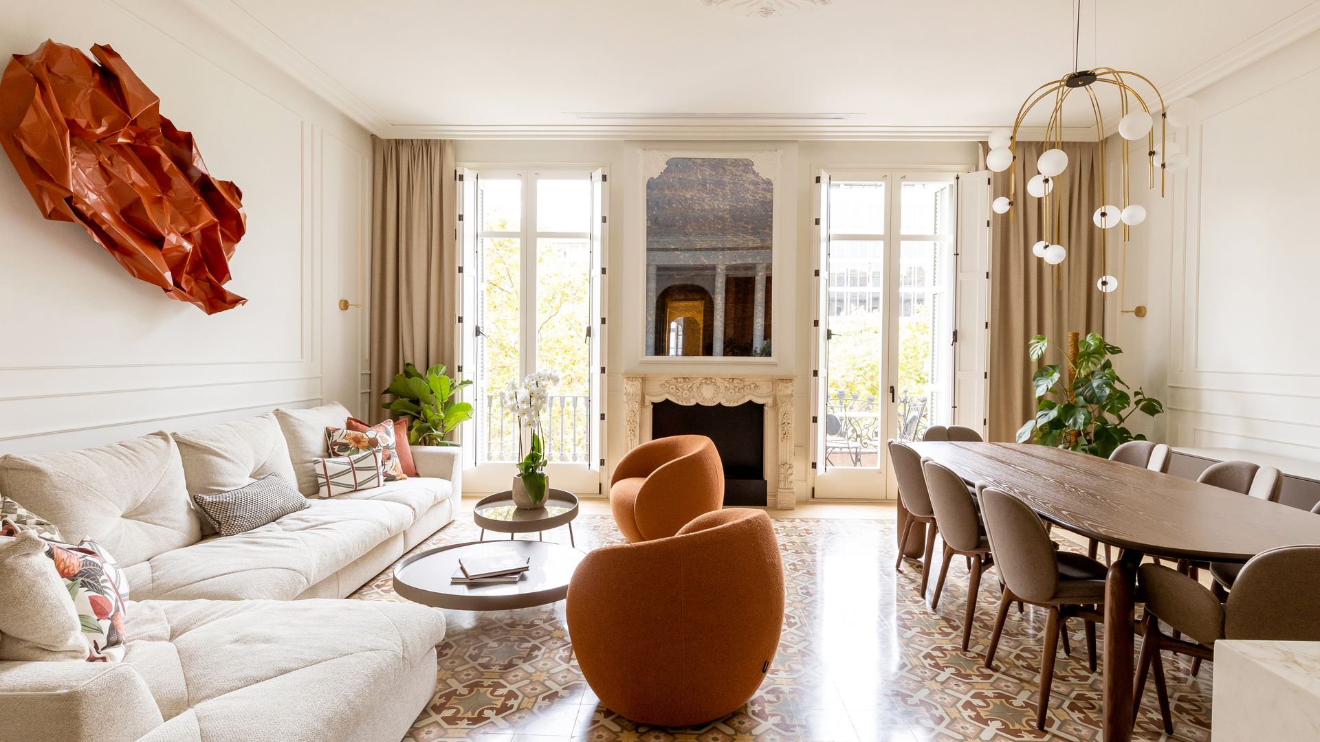 Salón comedor con chimenea, sofá blanco, escultura colgando en la pared del sofá, butacas naranjas, mesa de comedor ovalada con sillas de madera, lámpara de techo escultura 