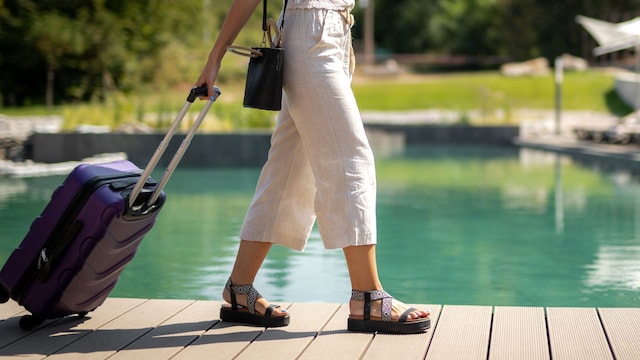 Mujer de cintura para abajo con pantalón de lino, sandalias de tiras y maleta entrando a un resort