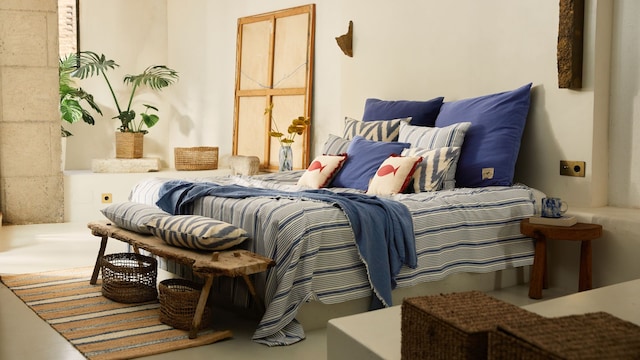 Dormitorio con ropa de cama de rayas, cojines azules y con motivos marineros, banco a los pies de madera, cestas y plantas