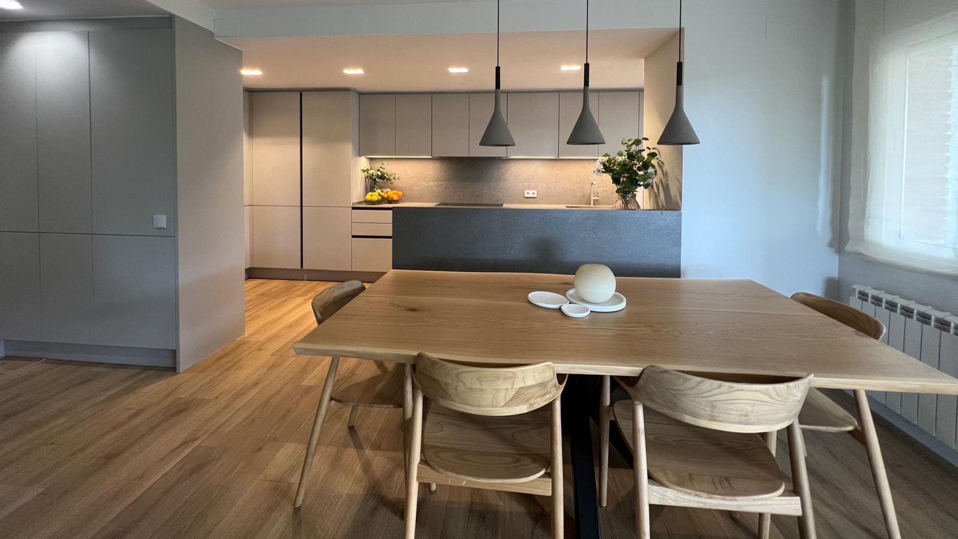 Cocina abierta al comedor, muebles grises, mesa de comedor de madera, suelo de madera, lámparas de techo 