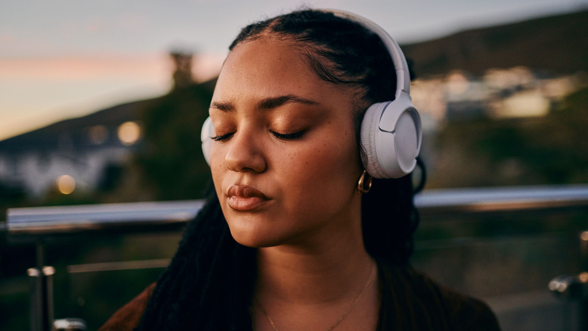 Mujer joven con los ojos cerrados escuchando música en la calle, con un anochecer de verano de fondo.