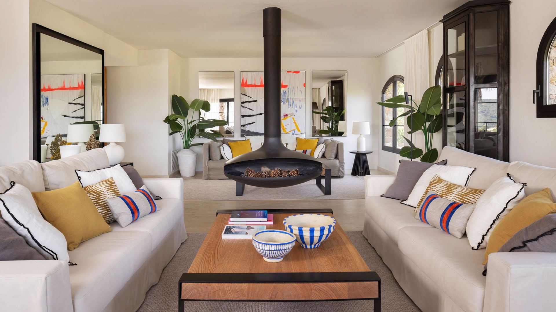 Salón con chimenea en el centro del espacio, sofás blancos, mesa de centro de madera con el marco y las patas negras, cojines en los sofás, plantas altas, vitrina negra