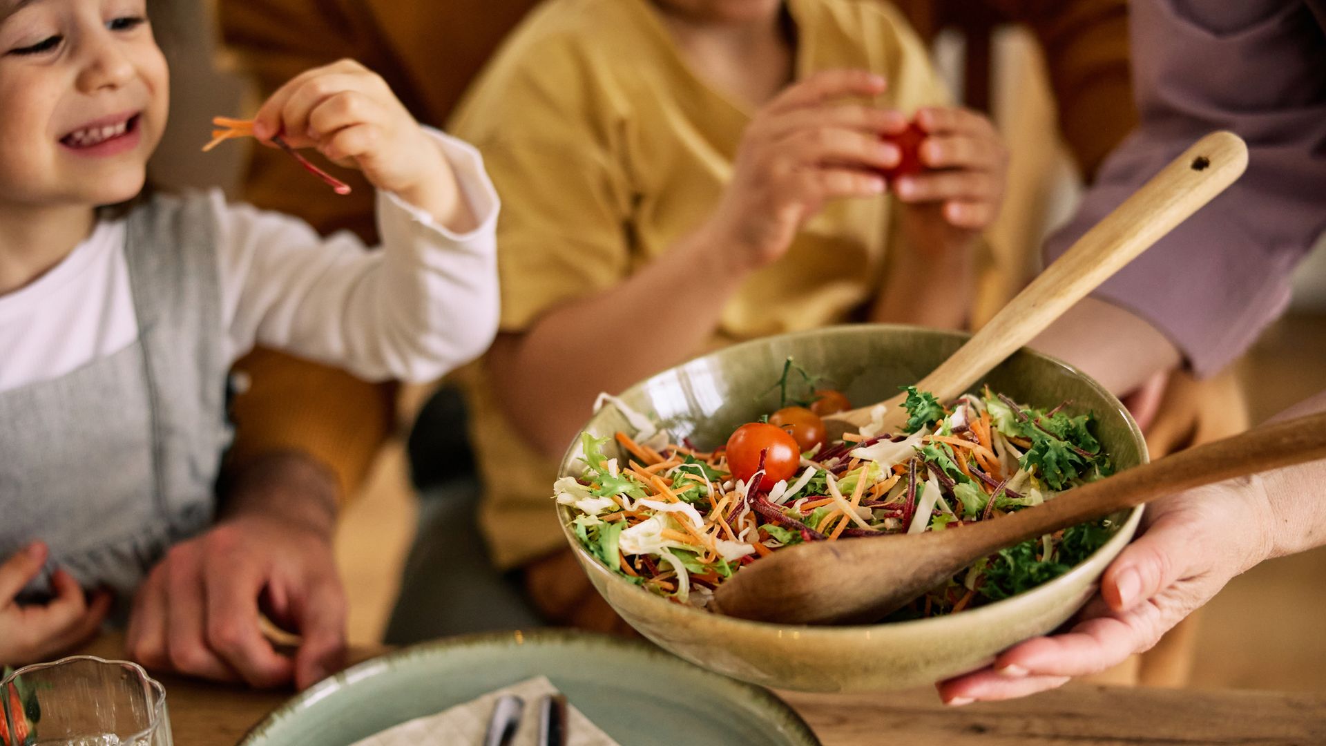 Niña sonriente comiendo un poco de ensalada con los dedos de una ensaladera que alguien sujeta en el centro de la mesa.