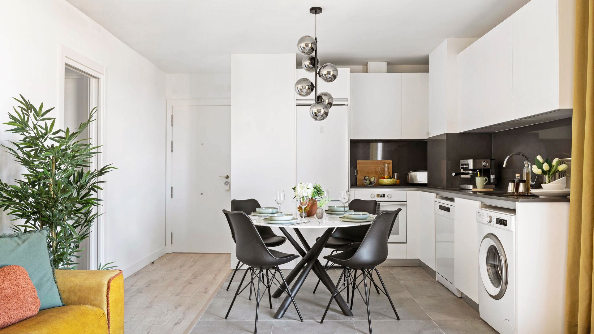Cocina abierta con muebles blancos hasta el techo, mesa redonda blanca, sillas negras, suelo porcelanico, lámpara de techo 