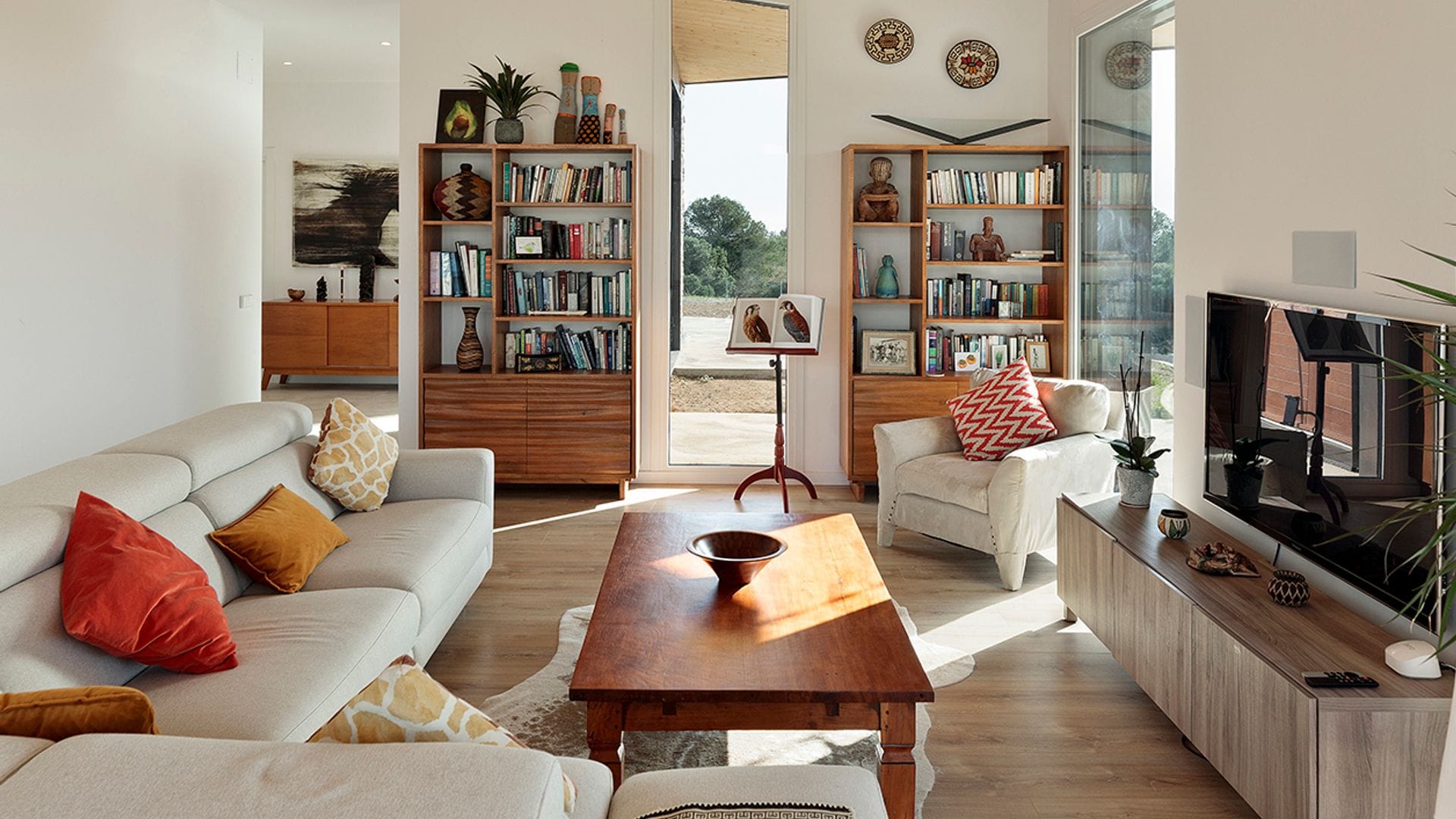 Salón con sofá blanco, grandes ventanales, mueble de televisión, mesa de centro de madera, suelo de madera, librerías de madera a ambos lados de puerta de salida al jardín 