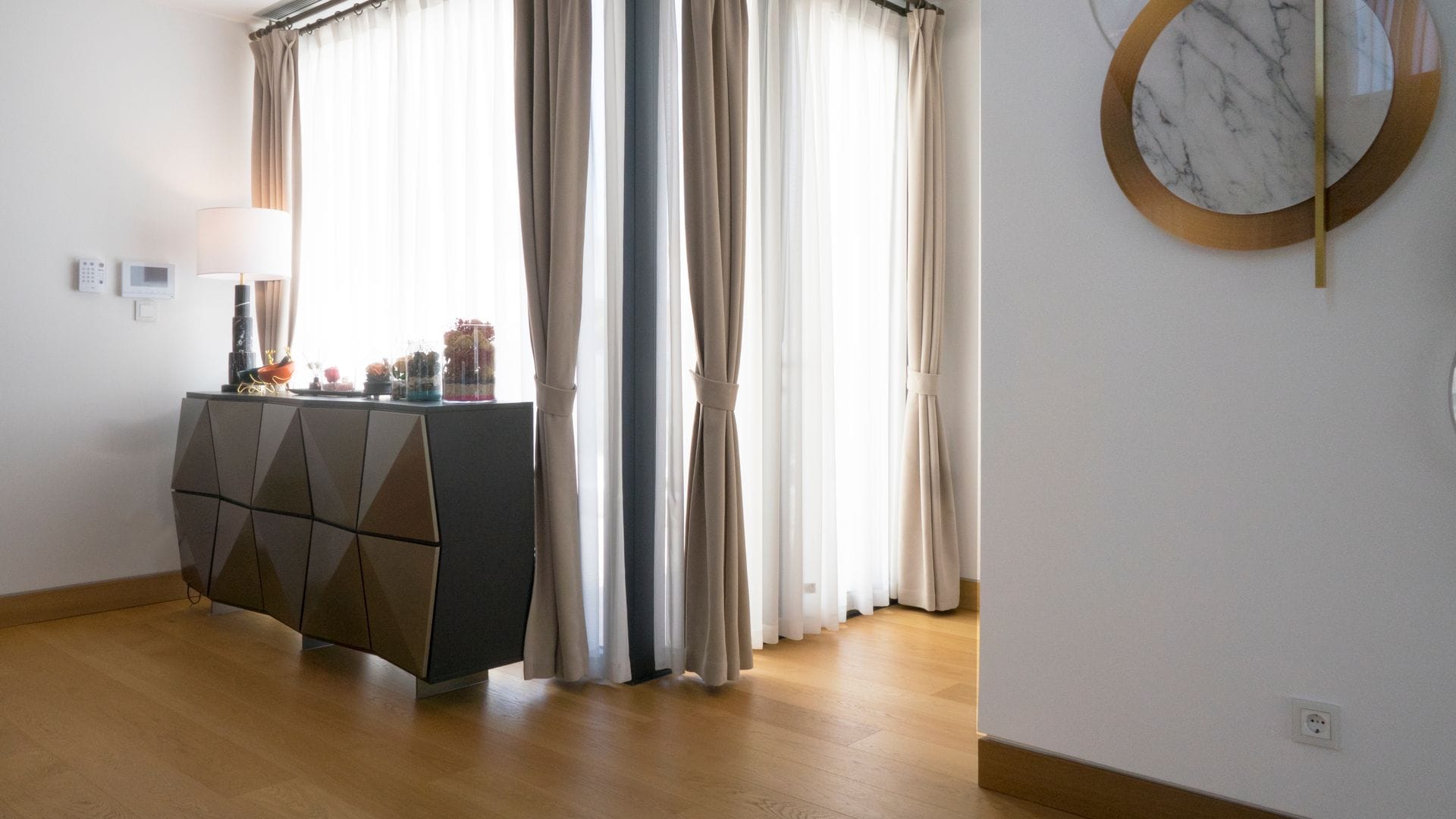 Recibidor con aparador moderno, lámpara de sobremesa, suelo de madera, ventanales con visillos y cortinas