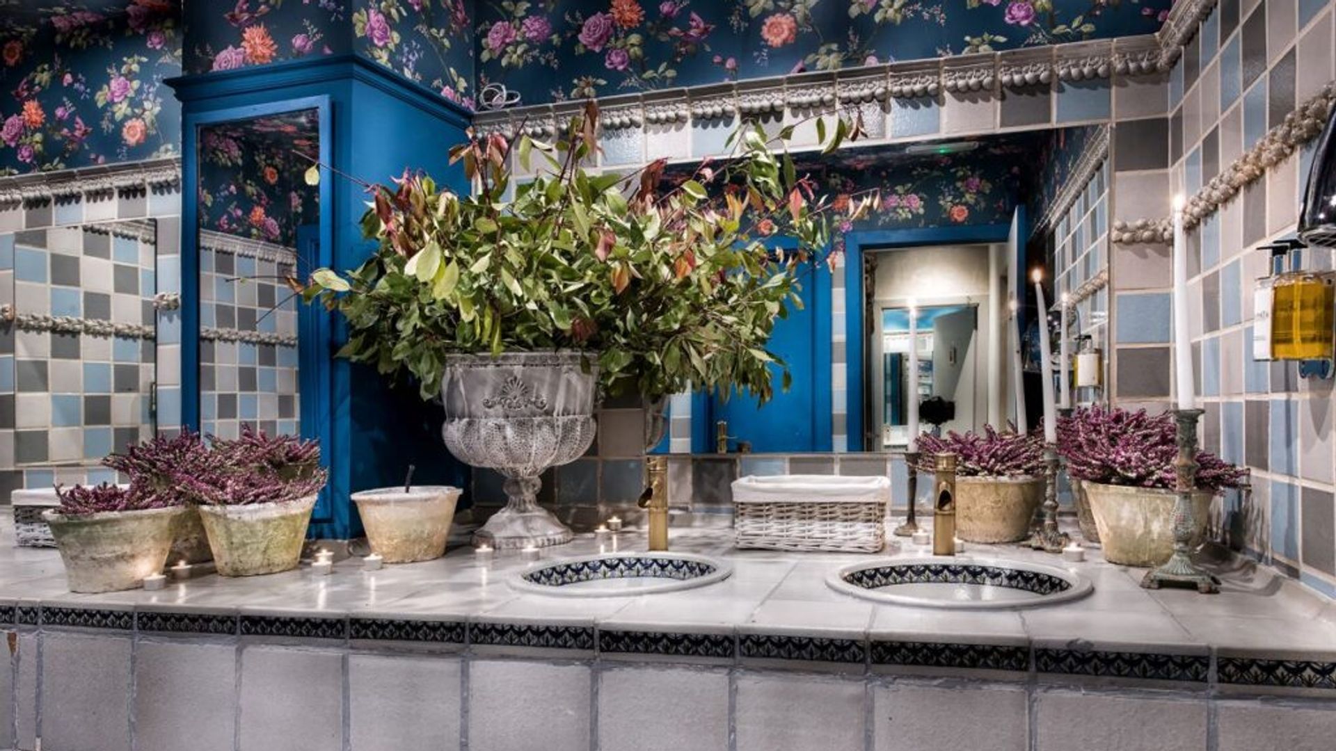 Baños con azulejos, encimera de azulejos grises, papel pintado azul oscuro con flores, espejo de pared a pared, jarrón con flores, lavabos 