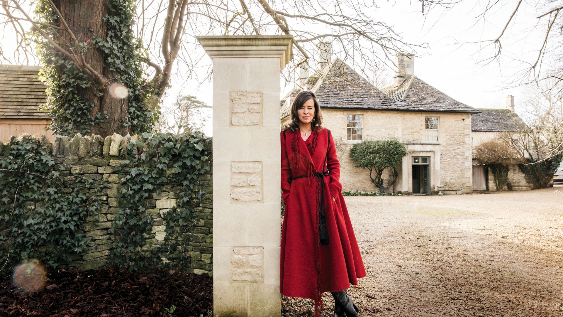 Jade Jagger, con abrigo rojo, se apoya en un pilar junto a una casa antigua.