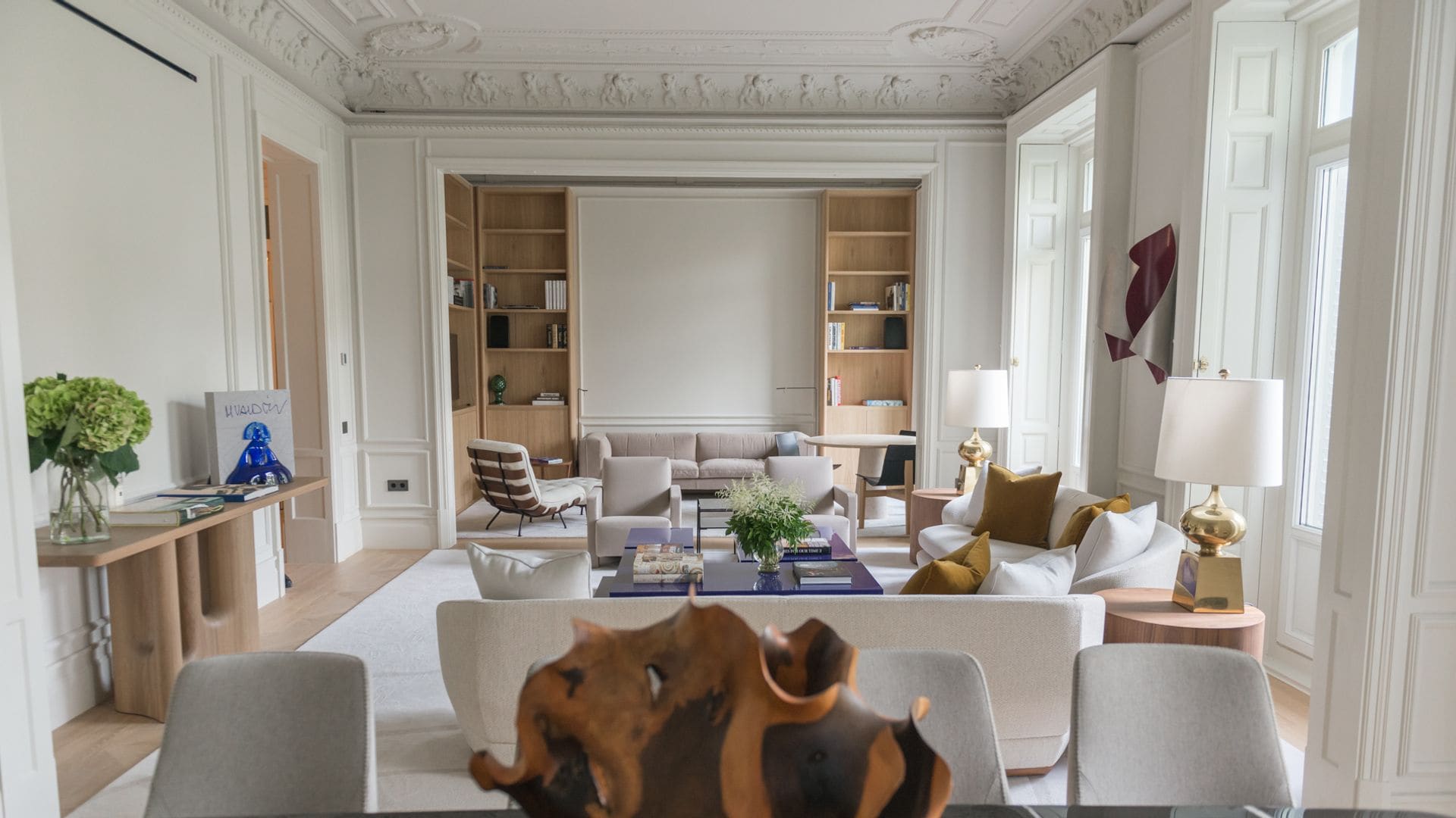 Salón comedor con molduras en el techo, sofás blancos, lámparas de sobremesa, librerías a medida de madera, consola de madera con jarrón con hortensias, libro adorno sobre la consola  