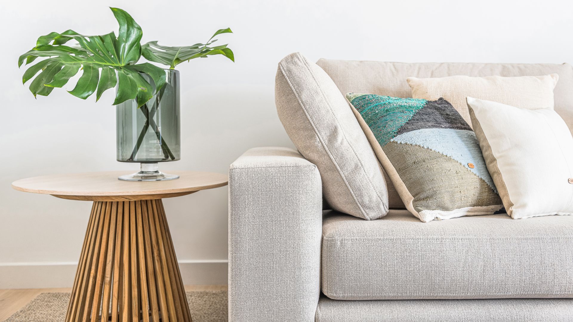Sofá beige con cojines en los mismos tonos, mesa auxiliar de madera con jarrón de cristal con hojas verdes