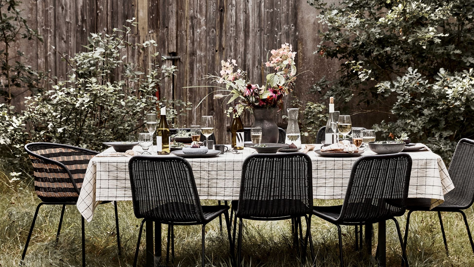 Mesa en el jardín con mantel de cuadros blanco, sillas negras, jarrón con flores