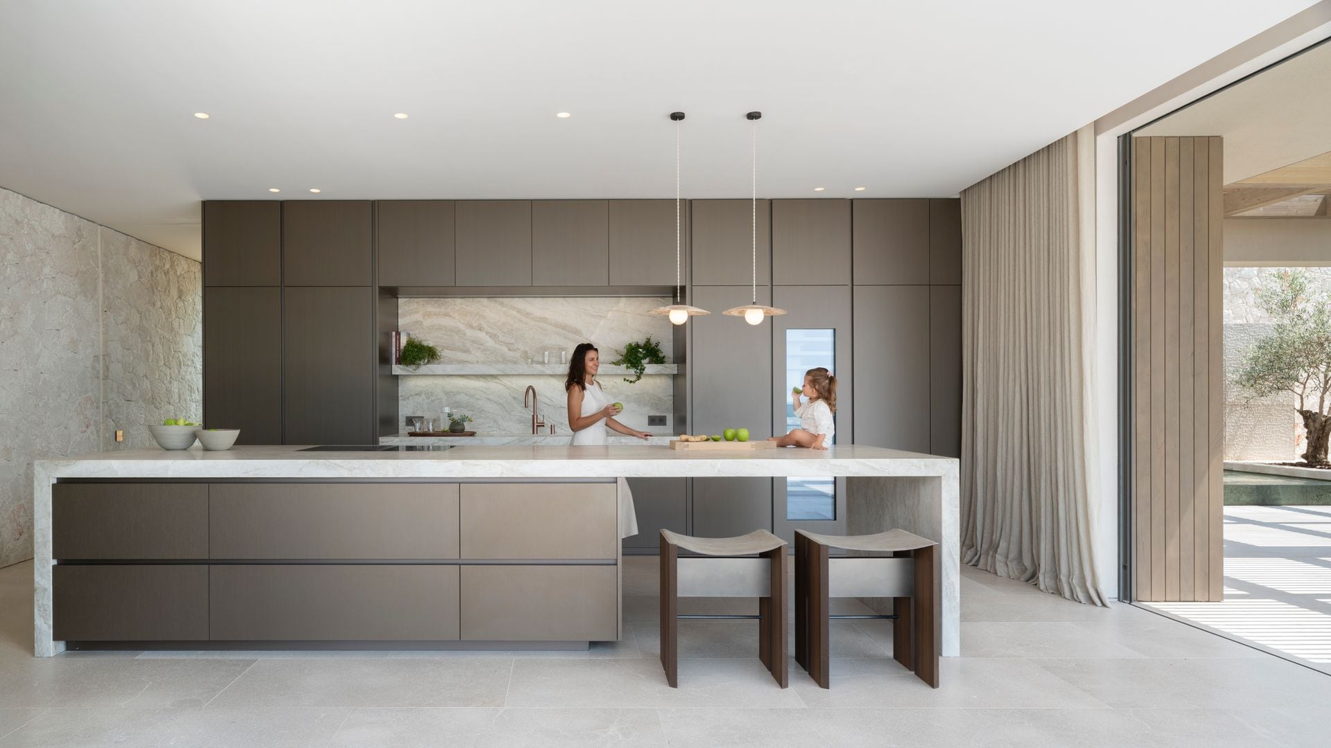 Cocina abierta al jardín con mujer y niña sobre la encimera, isla de cocina con zona para comer, muebles hasta el techo en color visón, lámparas de techo y focos empotrados