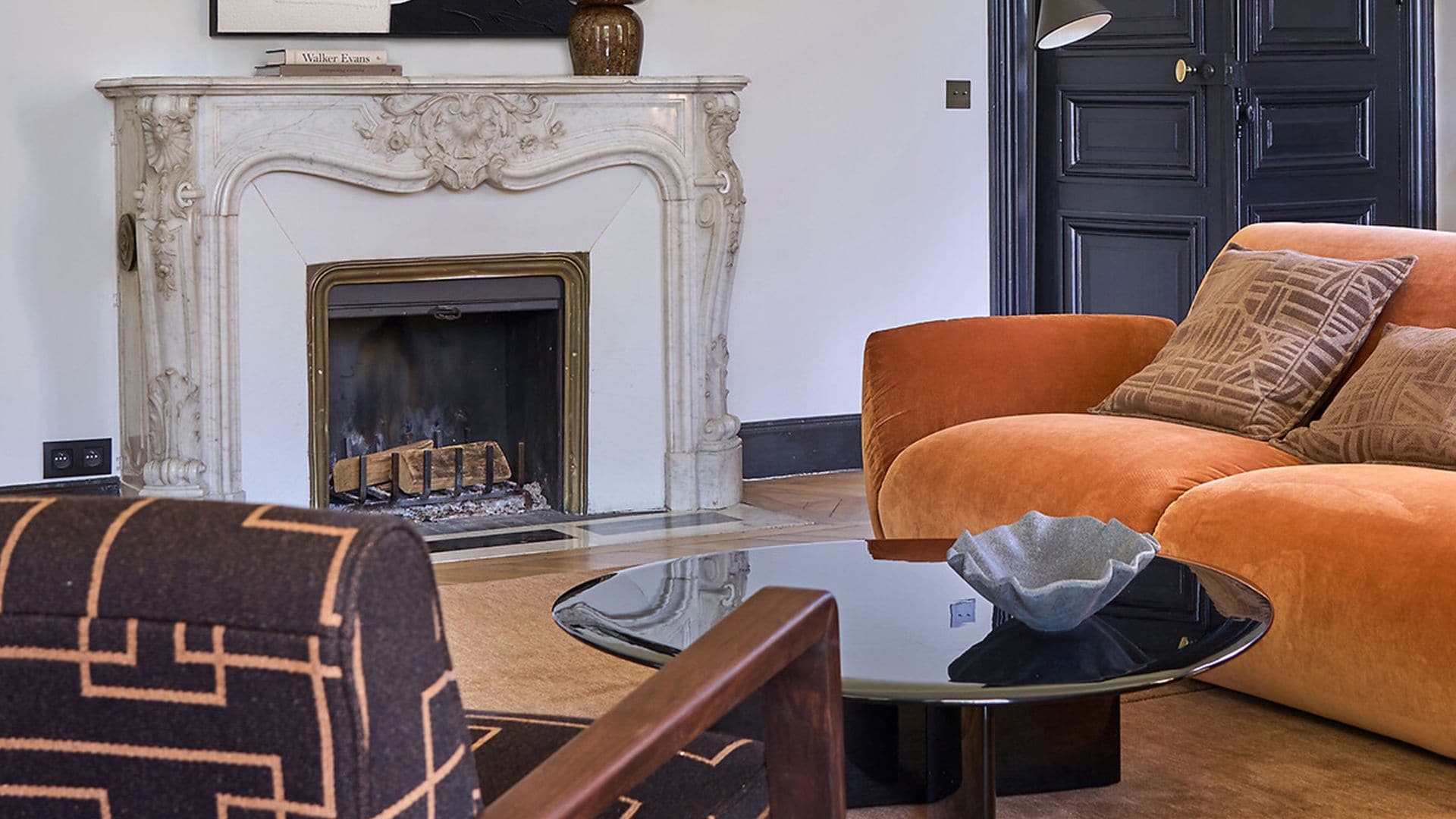 Salón con chimenea, cuadro sobre la chimenea, chimenea clásica, puertas de paso negras, mesa de centro negra brillante, sofá naranja y butaca marrón con estampado geométrico naranja
