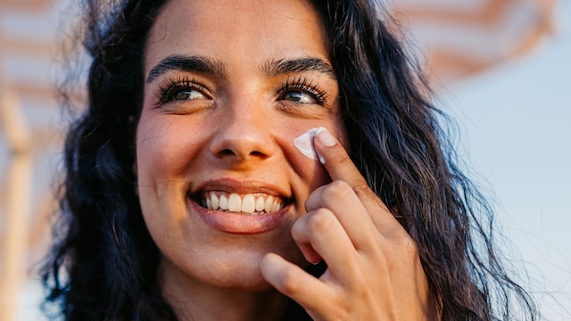 Mujer joven con pelo moreno rizado aplicándose crema solar en el rostro en la playa