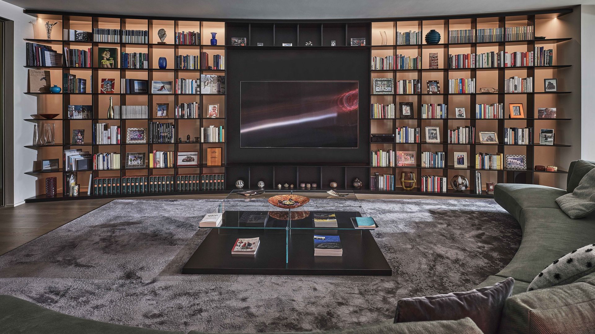 Salón con librería XL negra de suelo a techo y circular, television en el centro y libros, alfombra gris 