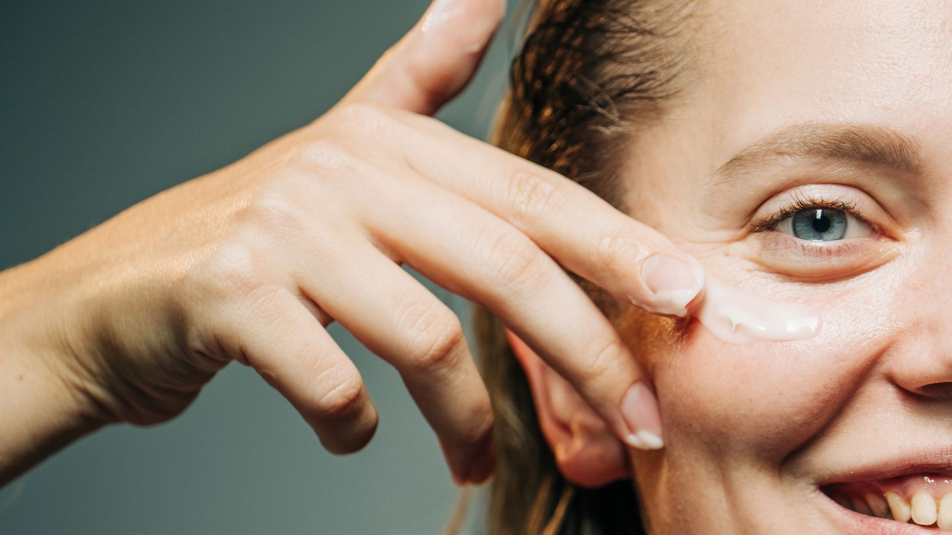 Mujer sonriente echándose crema en un lateral de la cara, en el pómulo bajo los ojos. Tiene los ojos azules y el pelo rubio.