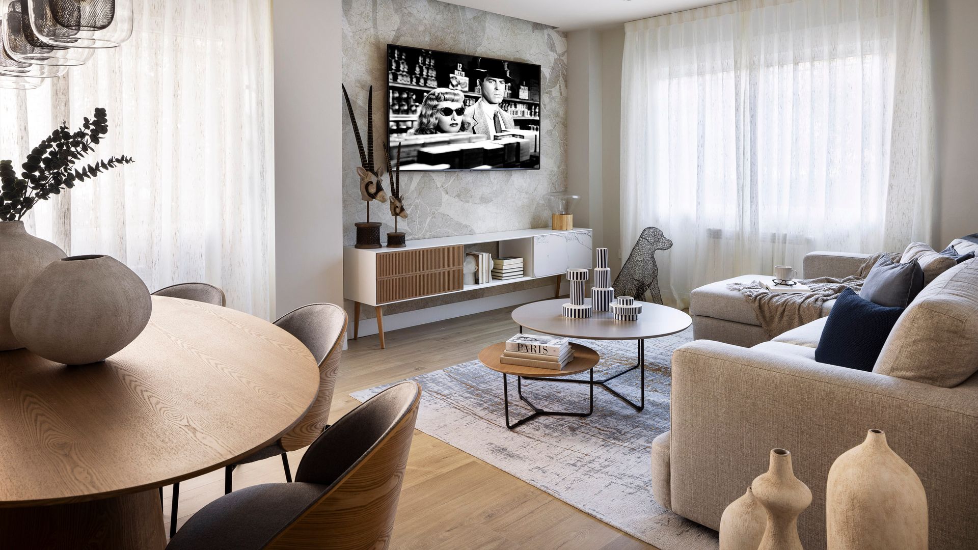 Salón comedor con televisión colgada en la pared, cortinas blancas, sofá beige, alfombra, mesa de centro redonda, mesa de comedor de madera, mueble de televisión