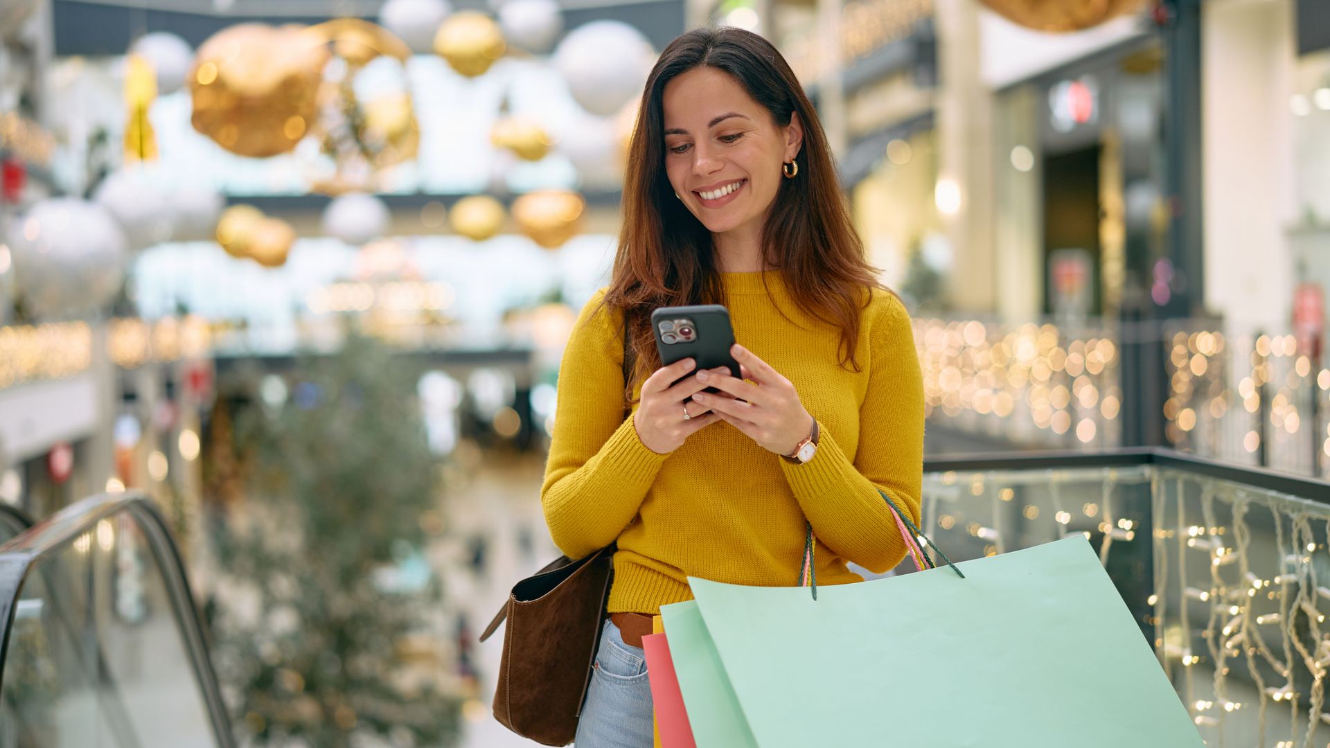 Mujer comprando en un centro comercial con bolsas de papel en el brazo y mirando sonriente al teléfono móvil. Lleva ropa de otoño-invierno