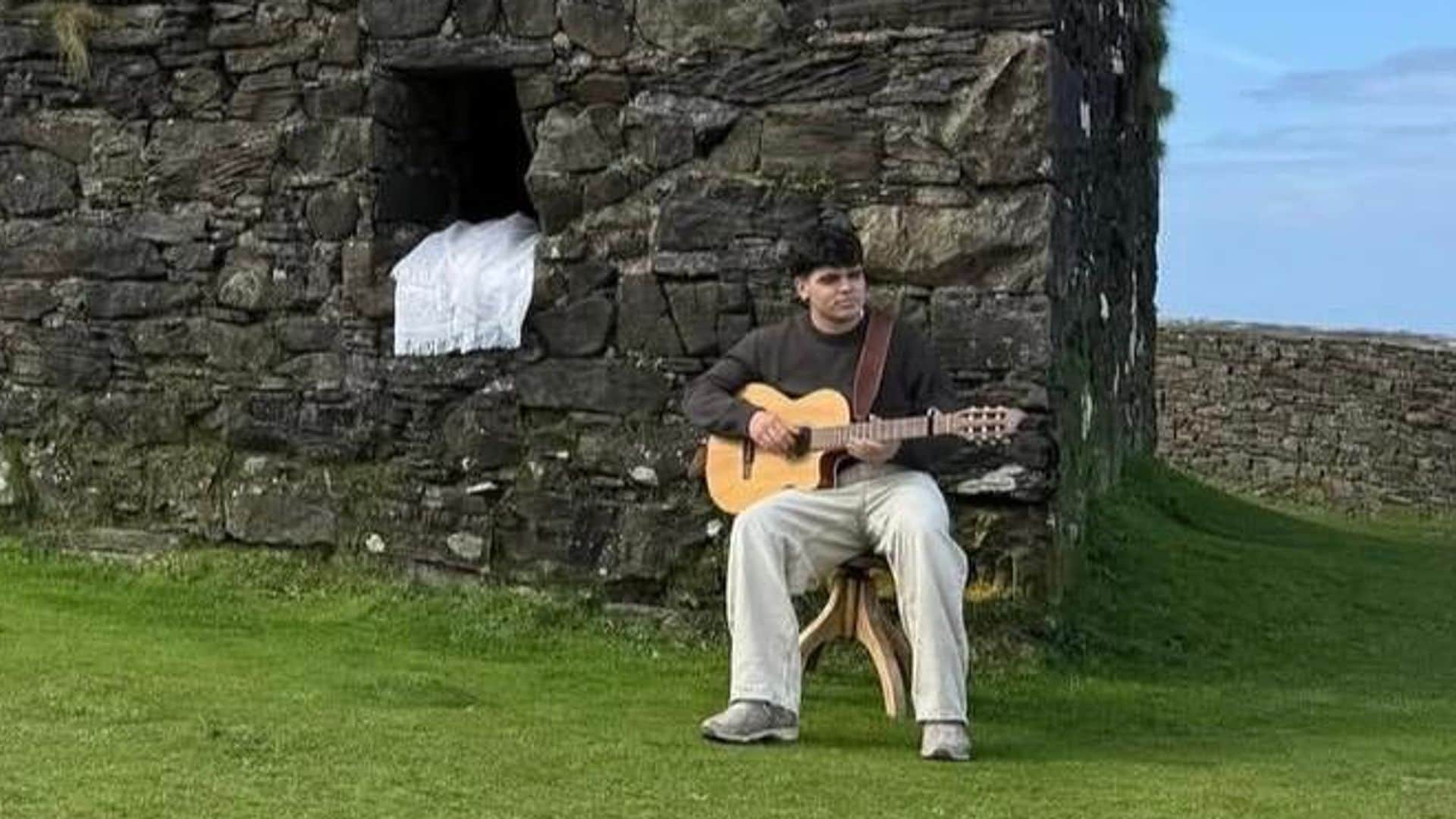 Iñigo Quintero etocando la guitarra en un prado verde junto a una casa de piedras