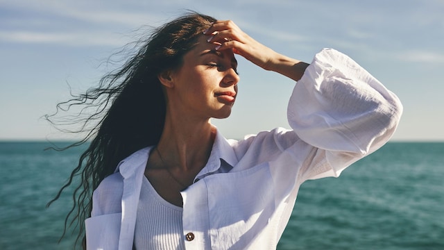 Mujer morena junto al mar con los ojos cerrados, el cabello al viento y la mano en su frente