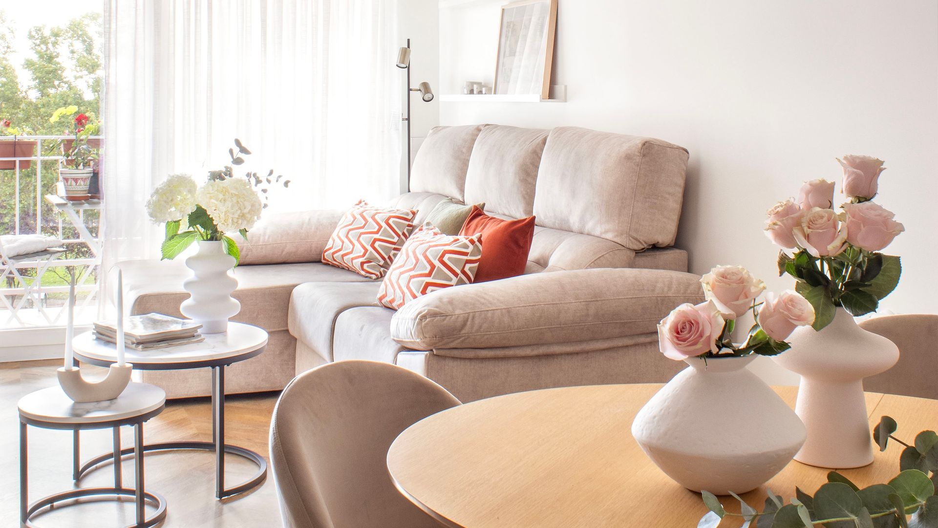 Salón comedor con sofá beige, dos mesas auxiliares cojines en color rojo con estampado geométrico, mesa de comedor de madera redonda, dos jarrones blancos con rosas de color rosa, cortinas blancas, ventanal con acceso a una terraza