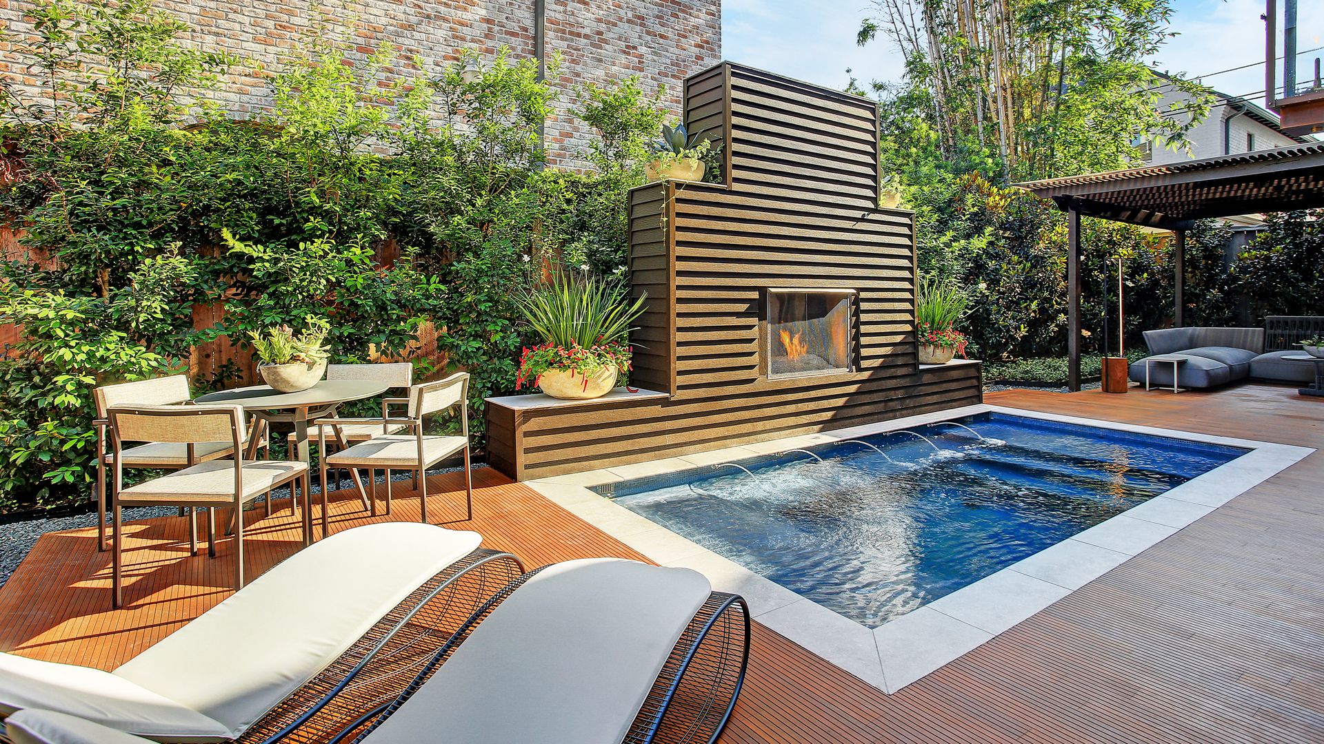Jardín con piscina, tumbonas, chimenea exterior, pérgola de madera, comedor con mesa y sillas, suelo de madera alrededor 