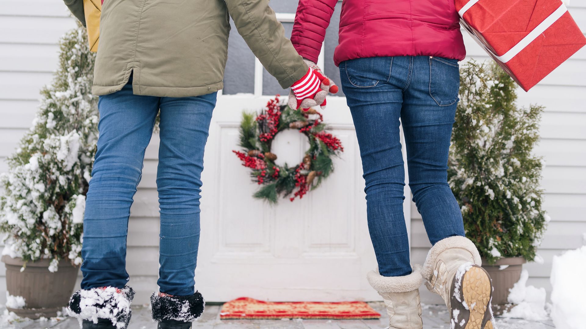 Madre e hija llegan a una casa de la mano con paquetes de Navidad. Imagen de ellas por detrás, con felpudo y corona de Navidad en la puerta. Ambiente nevado.