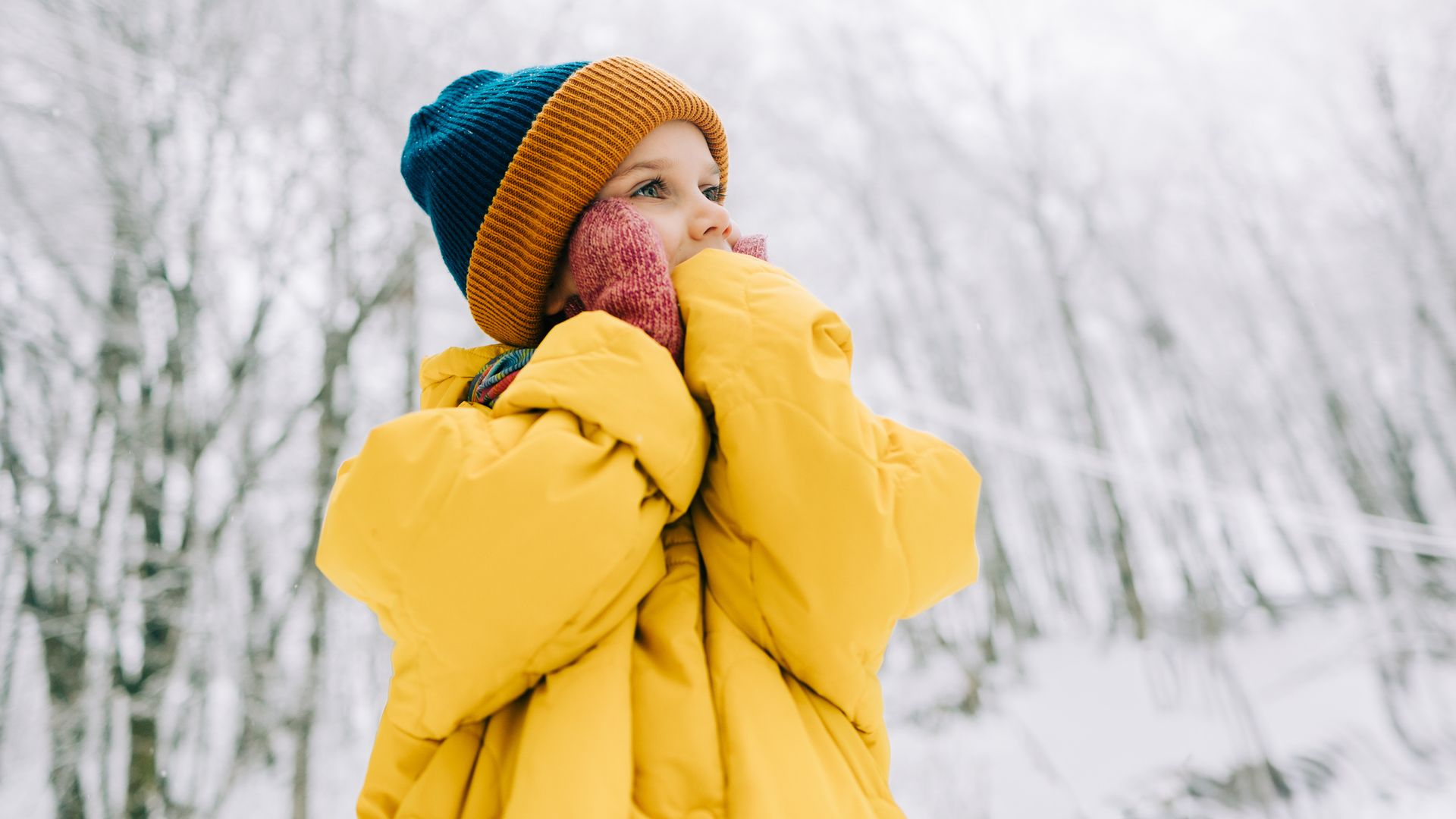 Niña sonriendo en la nieve bien abrigada en medio de la naturaleza. Lleva gorro de lana en azul y marrón, además de un abrigo amarillo y guantes rosas.