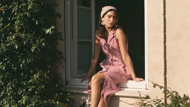 Mujer morena joven sentada en el alféizar de una ventana con vestido de cuadros vichy rojo y blanco