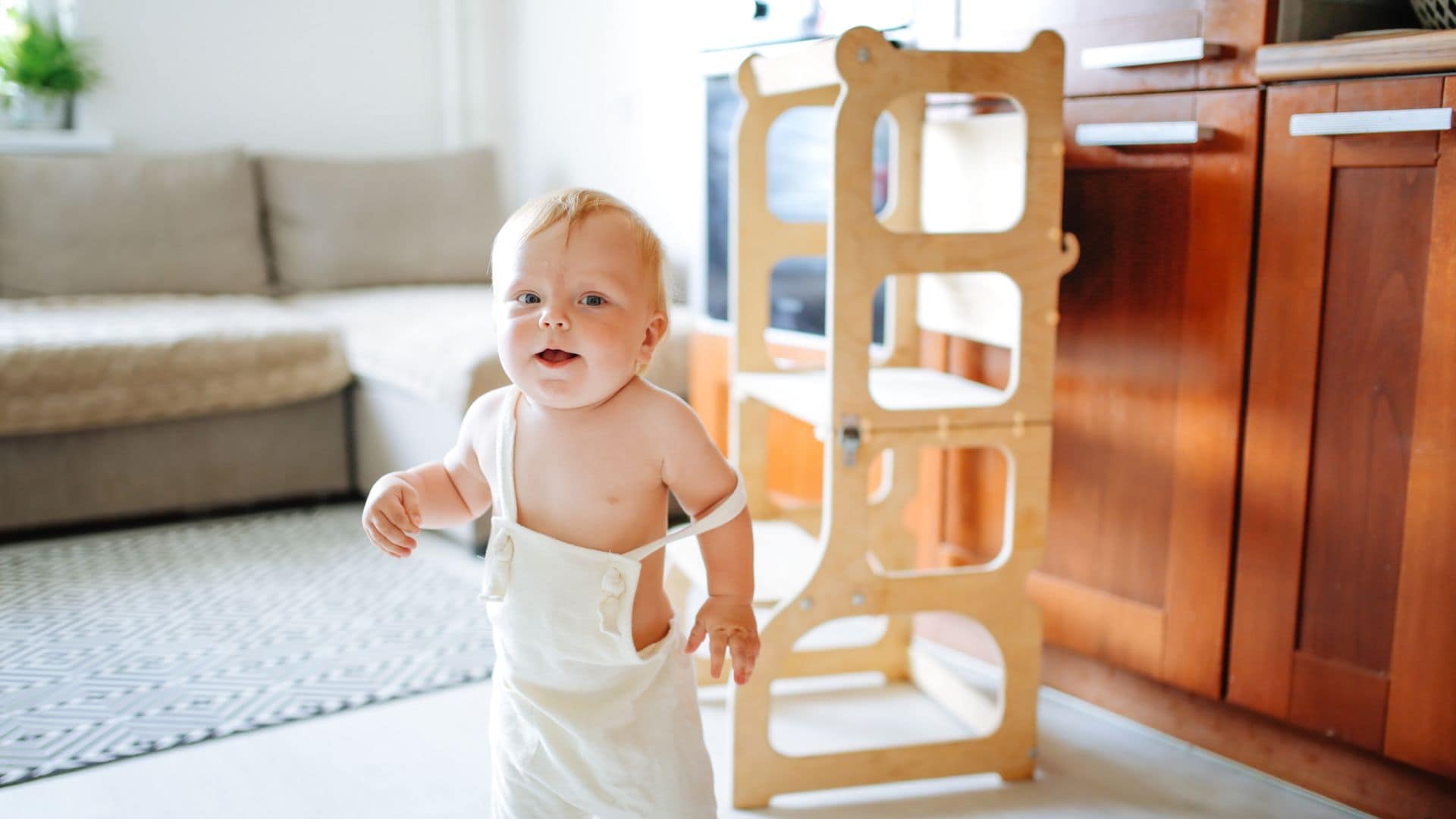 Niño bebe andando con una torre Montessori detrás de madera. El niño va con un peto beige y está sonriendo a la cámara.