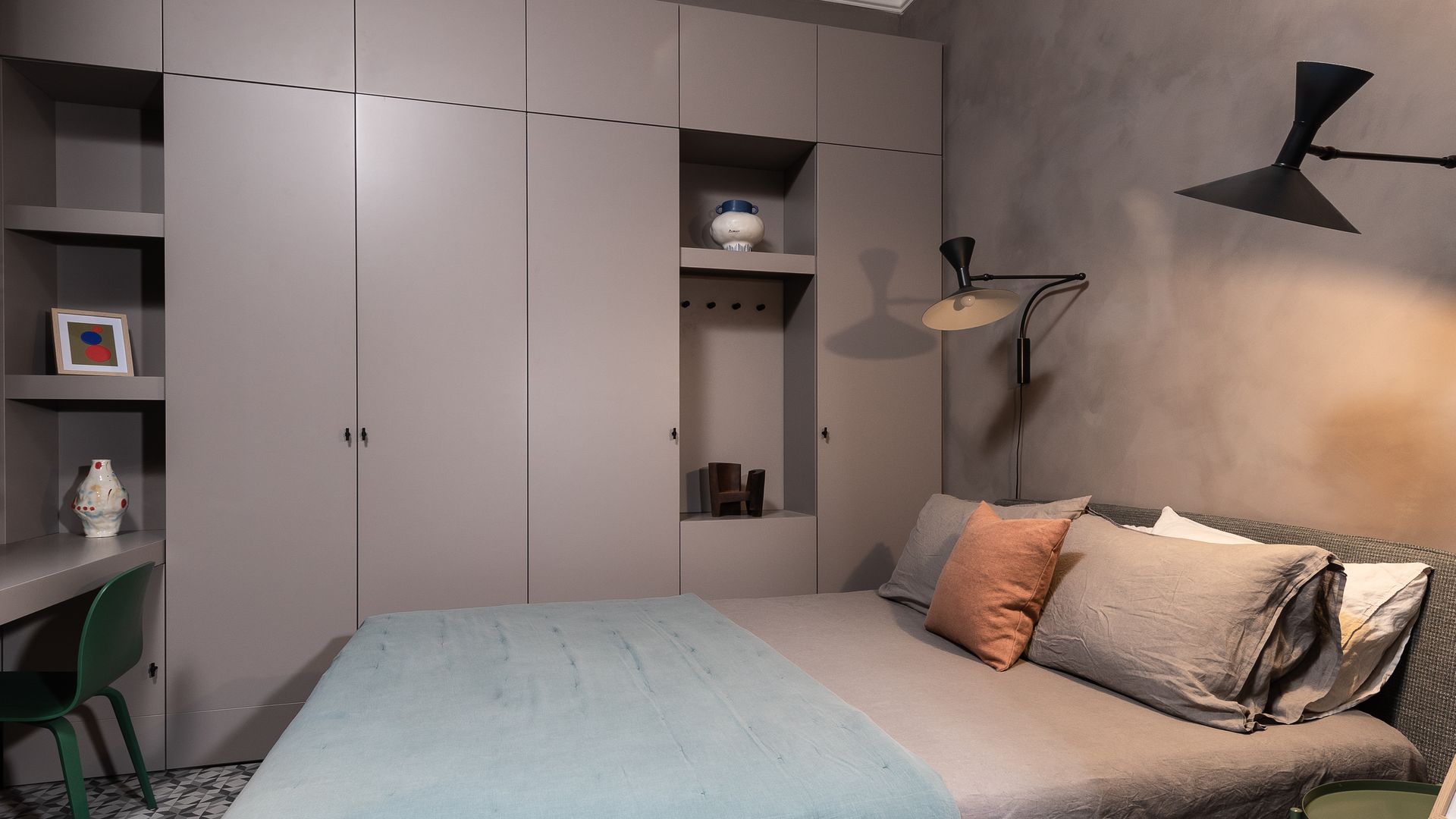 Dormitorio con paredes grises, alfombra, armario a medida gris, apliques de pared negros, ropa de cama gris y plaid azul, mesita de noche verde 