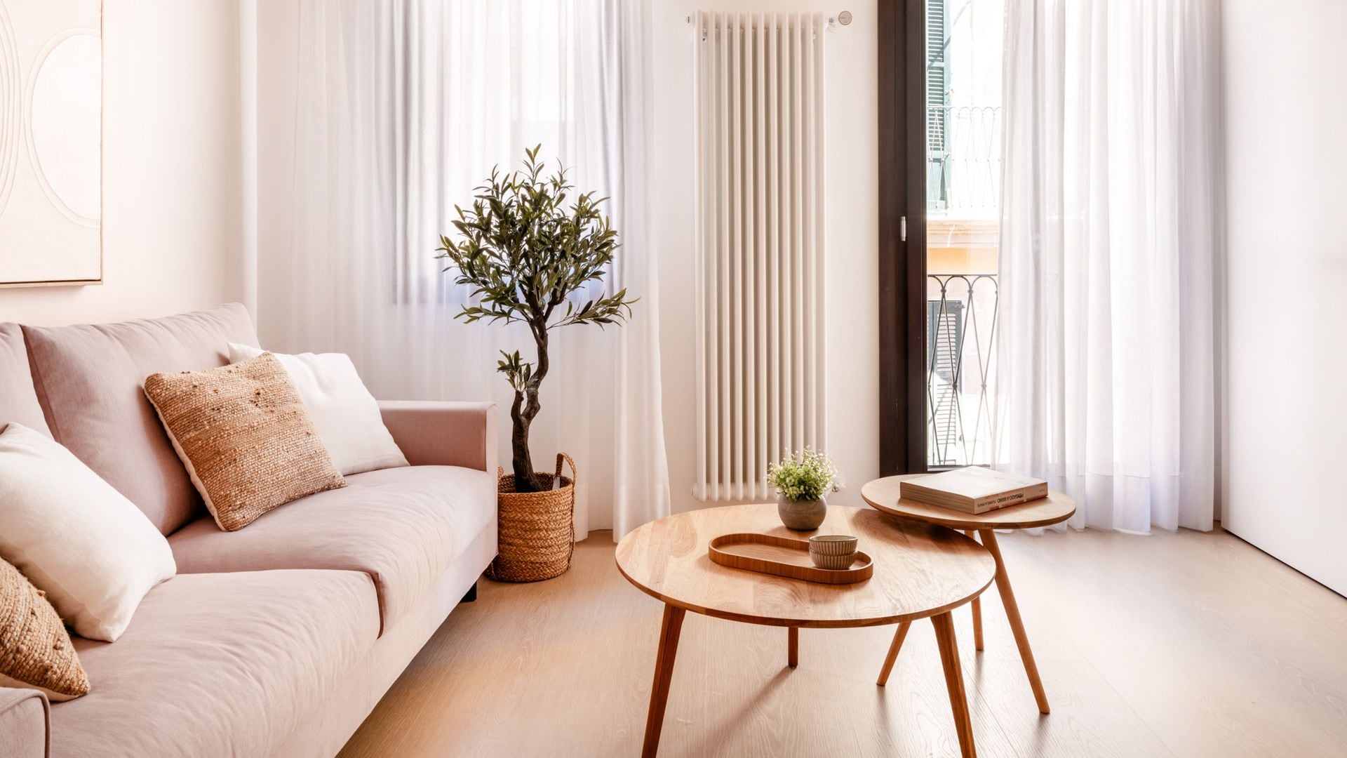 Salón con vigas de madera en el techo, ventanal con salida balcón, visillos blancos, suelo de madera, radiador XL, sofá rosa palo, mesas de centro de madera, planta en macetero de fibras vegetales